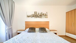 Un lit bien fait, recouvert d’une couvre-lit à motifs, avec une tête de lit, une table de chevet, une commode, une armoire et un tableau représentant un paysage urbain accroché au mur.