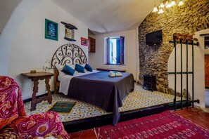 Une chambre avec un mur en pierre, un lit doté d’une tête de lit, une télévision, une fenêtre et une petite table.