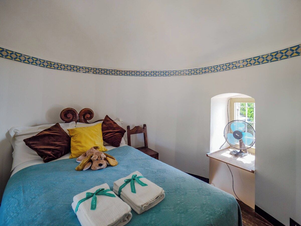 Un lit bien fait, avec une couverture bleue, des oreillers et un ours en peluche. Deux serviettes pliées, ornées de rubans verts, sont posées sur le lit. Une petite table avec un ventilateur se trouve à côté du lit.