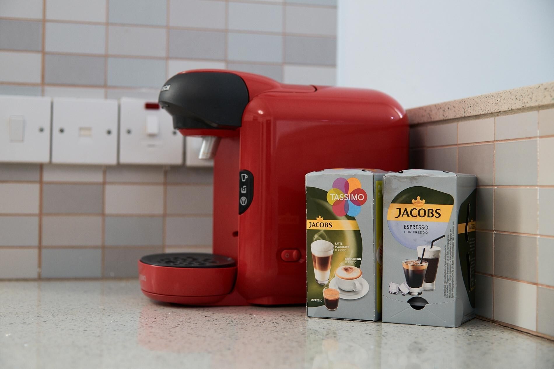 En röd kaffemaskin och två förpackningar med kaffekapslar, märkta med "TASSIMO" och "JACOBS", står på en köksbänk.