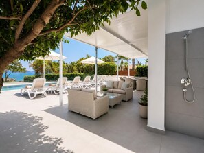 An outdoor lounge area with white lounge chairs, a sofa, and umbrellas, overlooking a pool and a clear sky.