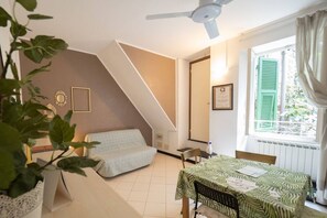 Une pièce avec un lit, une table recouverte d’une nappe, un ventilateur de plafond, une fenêtre avec des volets et une plante.