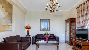 A living room with a brown leather sofa, a glass coffee table, a TV on a wooden stand, a chandelier, and a framed picture on the wall. - Villa Kalithea - Three Bedroom Villa, Sleeps 7 (Argostolion)