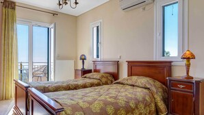 A bedroom with two beds, a bedside table with a lamp, a radiator, and a window with curtains. - Villa Kalithea - Three Bedroom Villa, Sleeps 7 (Argostolion)