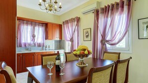 A dining area with a wooden table, chairs, a fruit bowl, wine glasses, and a chandelier. - Villa Kalithea - Three Bedroom Villa, Sleeps 7 (Argostolion)
