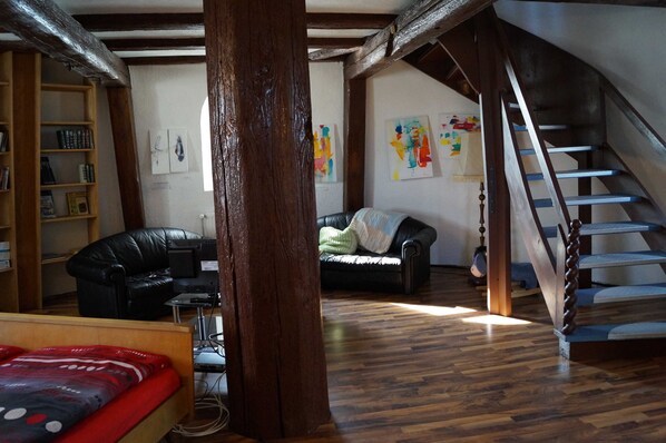A spacious living room with a wooden staircase, a black leather sofa, a wooden bookshelf, and a green blanket on a couch.