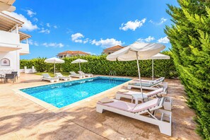 A swimming pool with lounge chairs and umbrellas, surrounded by greenery and buildings.