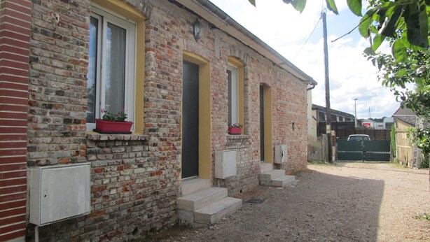 A two-story brick building with a yellow door and windows, a gravel driveway, and a gated entrance. - Gîte Côte Fleurie Lisieux center with parking (Lisieux)