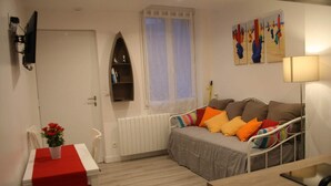 A bedroom with a bed, a bedside table, a radiator, a window with curtains, a wall-mounted shelf, and a painting. - Gîte Côte Fleurie Lisieux center with parking (Lisieux)