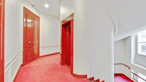 Un pasillo con alfombra roja, paredes blancas y una puerta roja.
