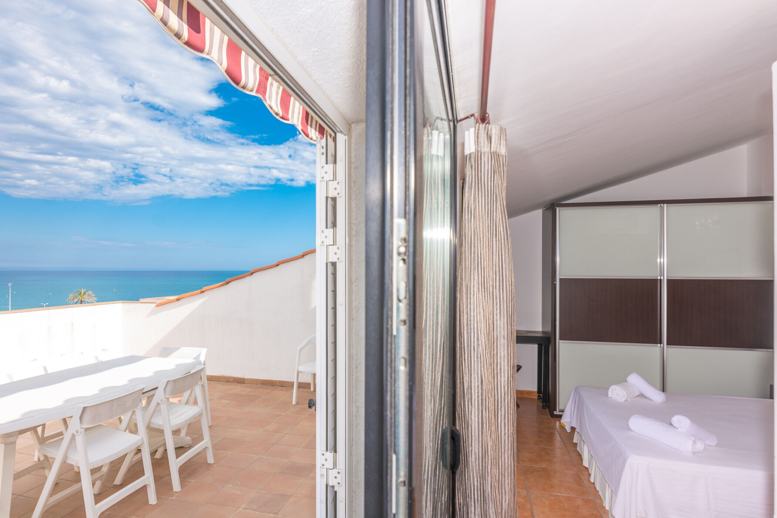 Een kamer met balkon en uitzicht op zee, een bed met witte lakens en een kleine tafel met stoelen.
