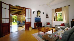 A spacious living room with a sofa, a television, and a coffee table. There is a large window with curtains and a view of greenery outside.