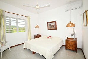 Quarto com cama, cadeira, mesa de cabeceira, luminária, quadro na parede e janela com cortinas.