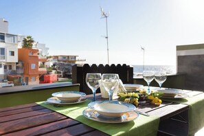 A table set for two with plates, glasses, and a small bouquet of flowers, overlooking a coastal area with buildings and a clear sky.
