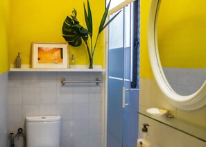 Ein Badezimmer mit Toilette, Waschbecken und Spiegel. An der Wand steht eine Pflanze und über dem Waschbecken hängt ein Bild in einem Rahmen.