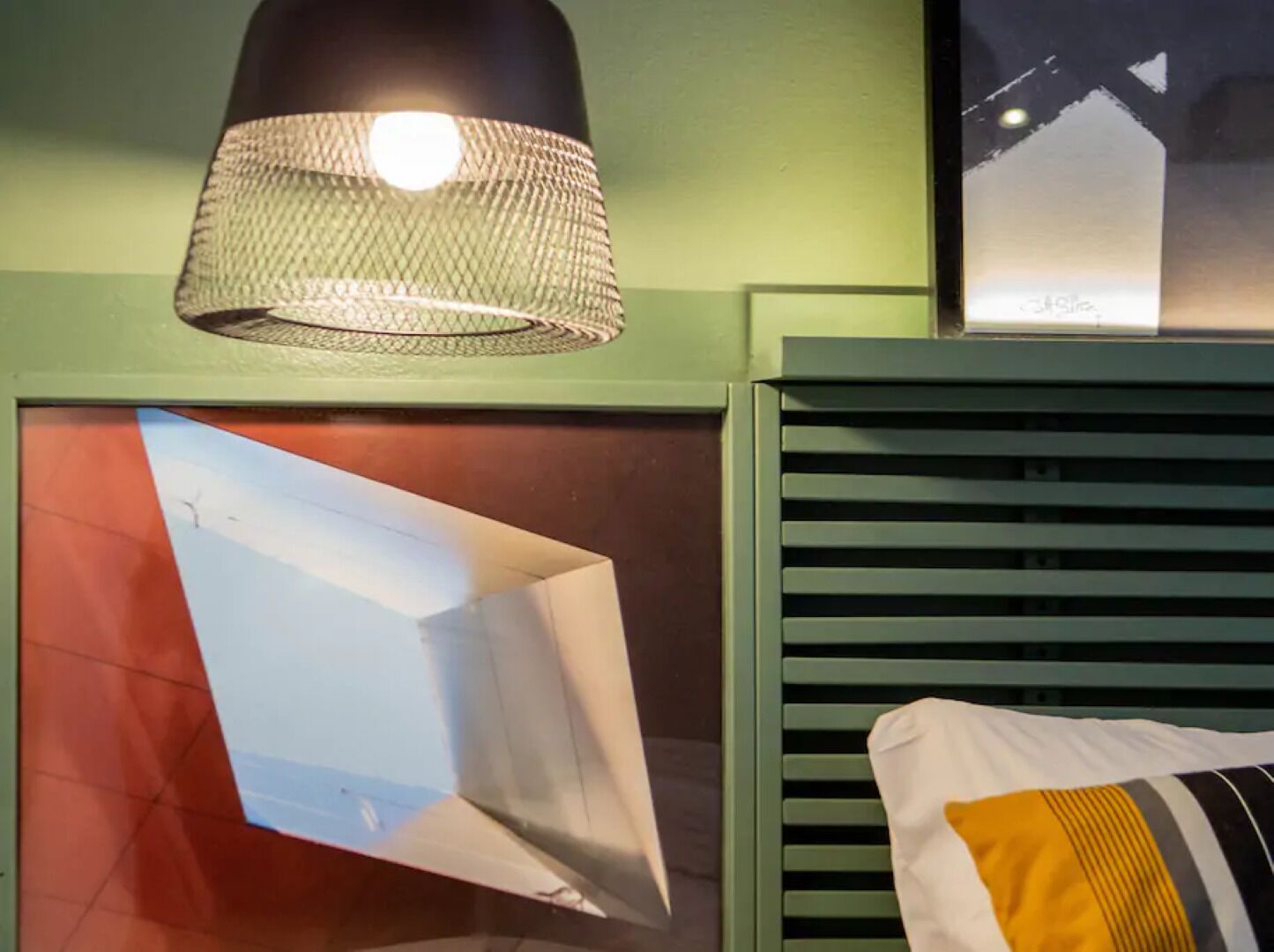 A room with a green wall, a framed picture, a hanging lamp, and a pillow with a striped cushion.