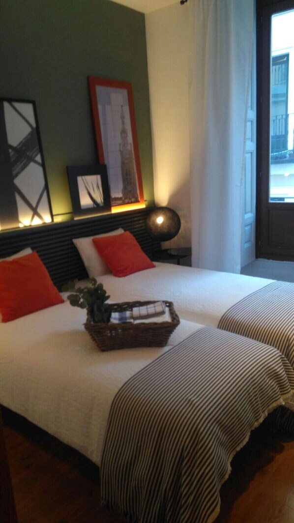 Habitación de hotel con dos camas, una mesita de noche con lámpara, un cuadro en la pared y una ventana con vista al exterior.