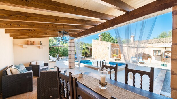 Un patio exterior con mobiliario de mimbre, una mesa de comedor con velas y vistas a una piscina y edificios.