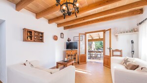 Una sala de estar con dos sofás blancos, un mueble de madera, un televisor y un comedor visible a través de una puerta abierta.