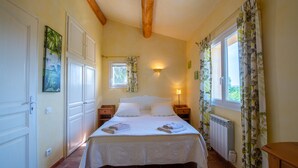 A bedroom with a bed, two bedside tables, a nightstand, a window with curtains, a wall-mounted light, and a radiator. -  The Vignaux bastide (Grimaud)