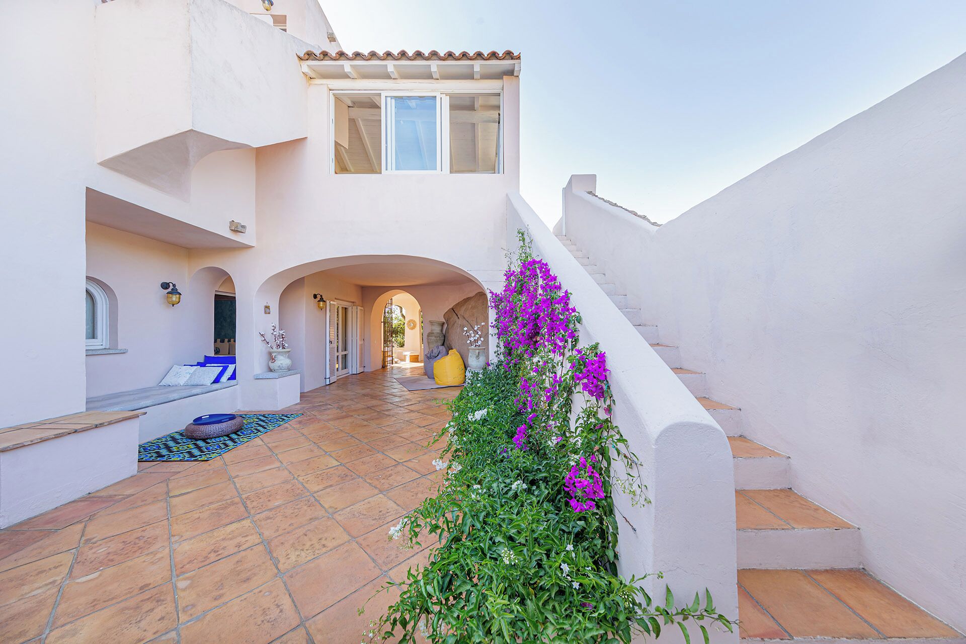 A courtyard with a tiled floor, arches, a staircase, and a vibrant purple flowering plant.