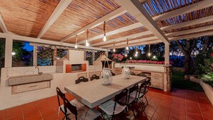 Un espace cuisine extérieur doté d’un plan de travail en pierre, de chaises en bois et de luminaires suspendus.
