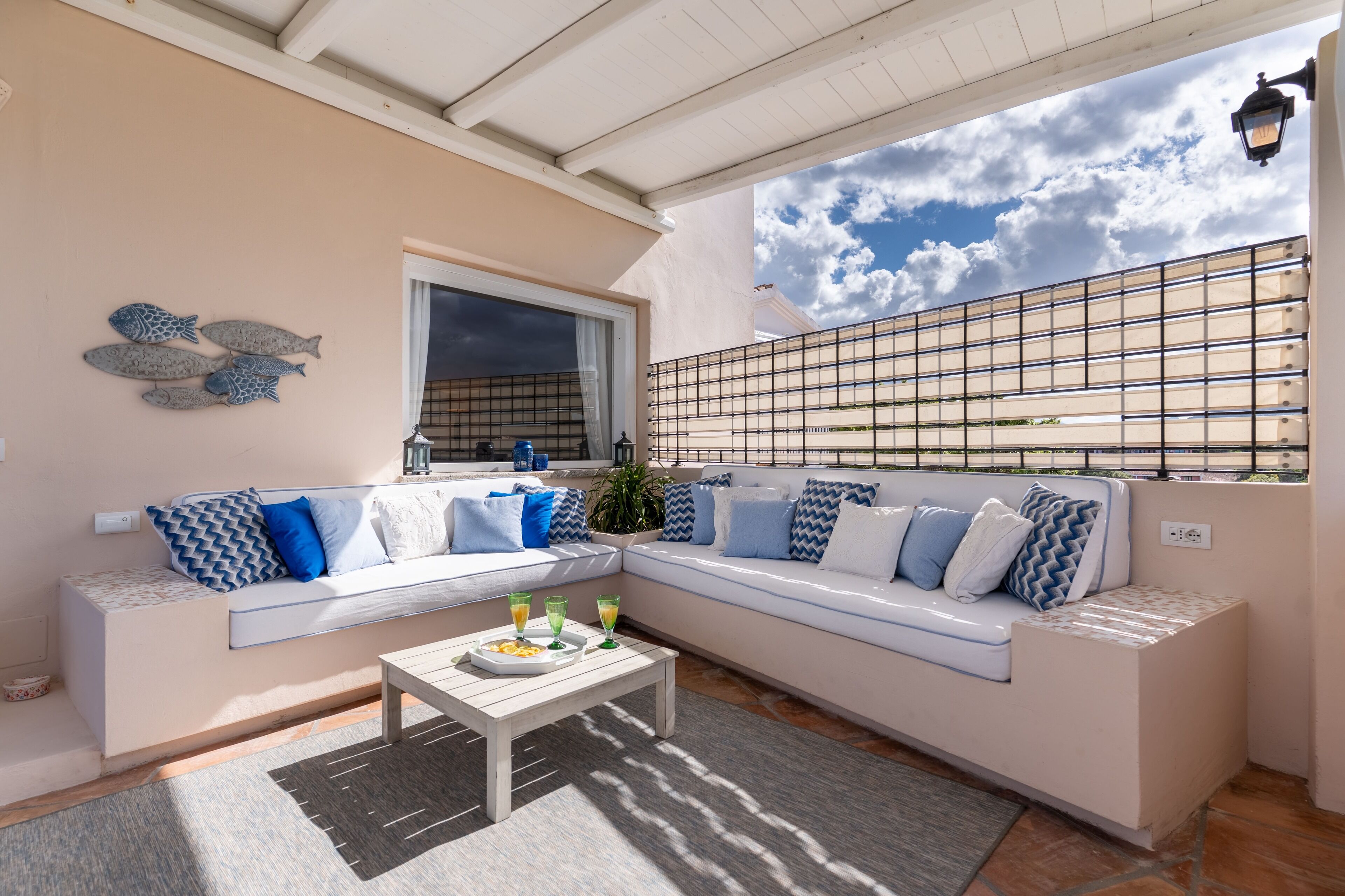 Un espacio al aire libre iluminado por el sol con un sofá blanco, cojines azules y blancos, una mesa de centro de madera con bebidas y una pieza decorativa de arte mural con peces.