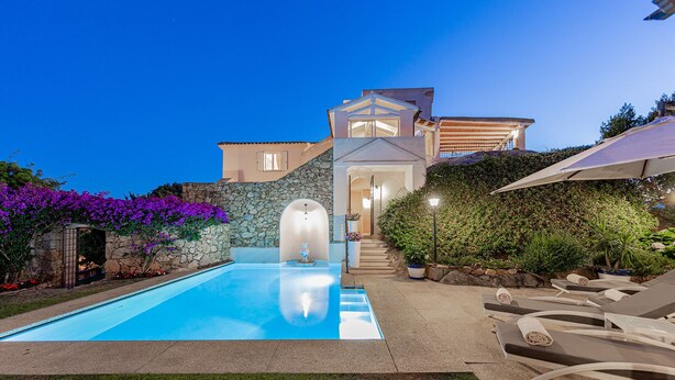 Une piscine bordée d’un mur en pierre, avec une maison moderne en arrière-plan.