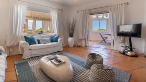 Un salon moderne avec un canapé blanc, une table basse et un téléviseur. On y trouve des rideaux, un vase avec des branches et une vue sur l’extérieur.