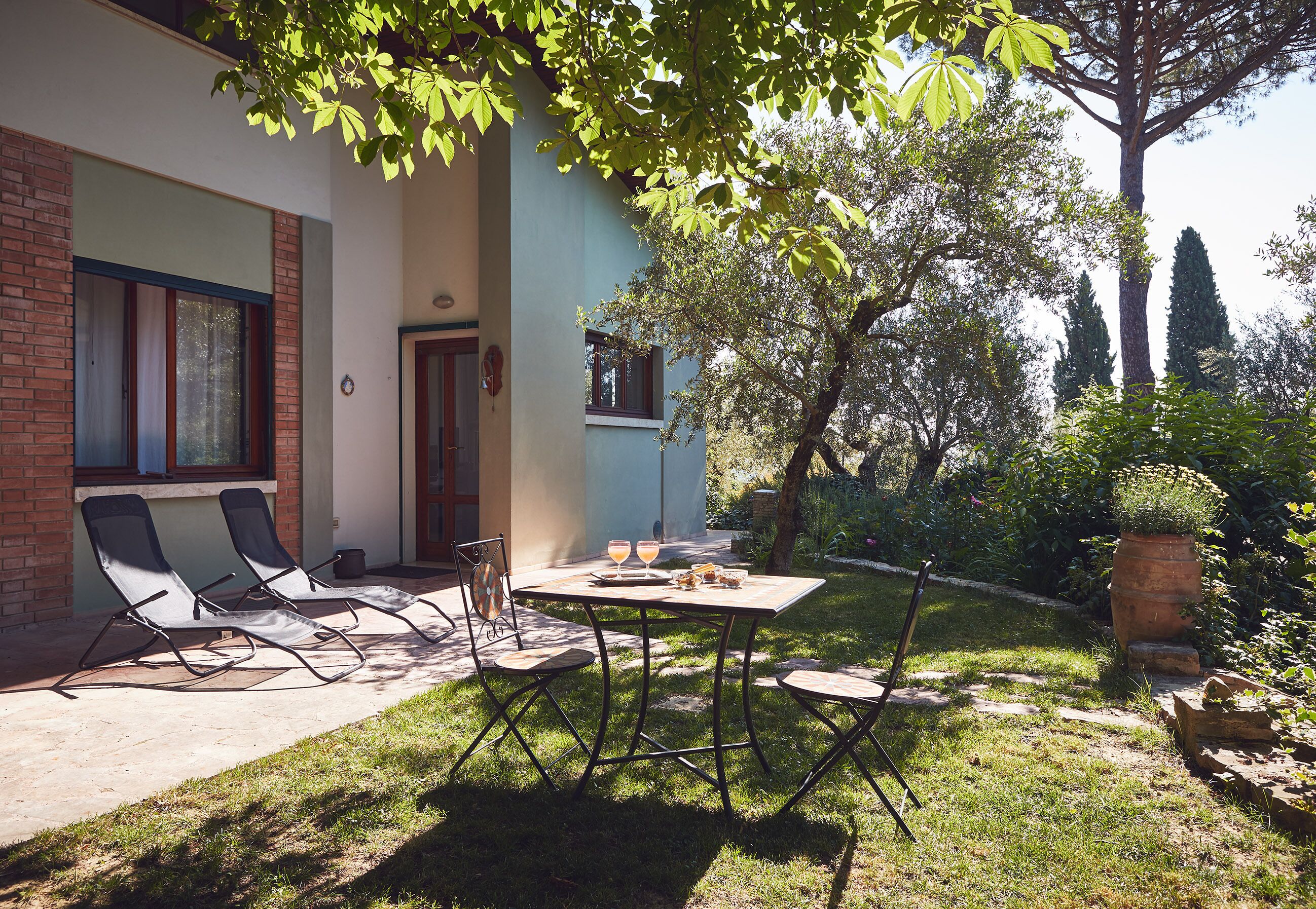 A patio with two lounge chairs and a table, surrounded by a garden with trees and a brick wall.