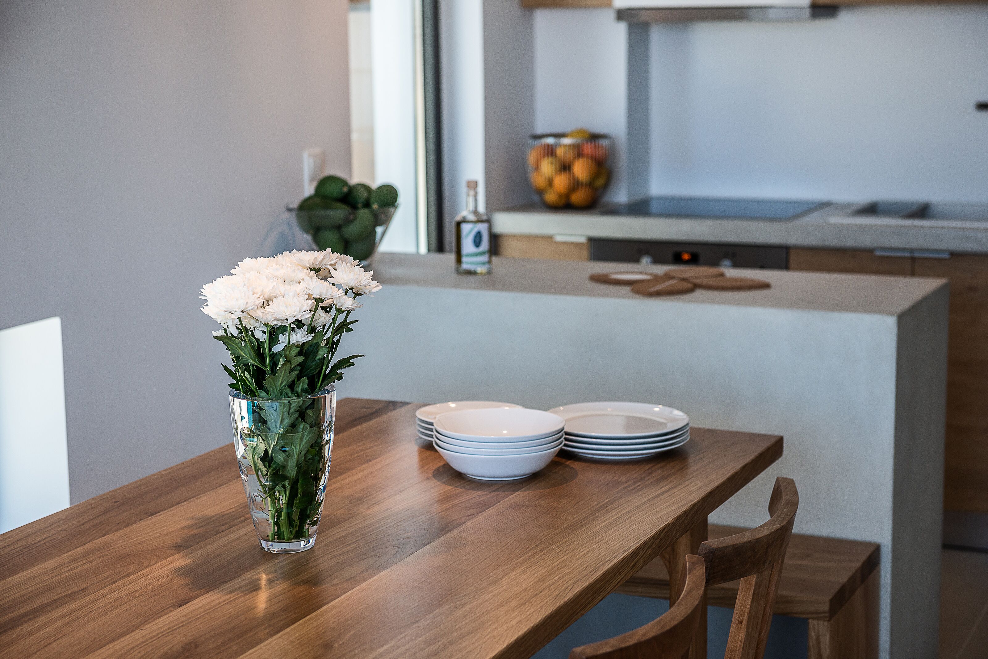 Una mesa de comedor de madera con un florero de vidrio transparente con flores blancas, platos blancos y un cuenco de fruta al fondo.