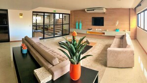 A modern living room with a sofa, a coffee table, and a potted plant. There is a staircase leading to another level, a flat-screen TV mounted on the wall, and a window providing natural light.