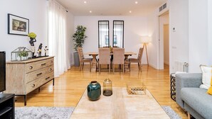 A modern living room with a wooden floor, a grey sofa, a dining table with chairs, a TV, and decorative items. - Deluxe Apartment Juan Bravo Suites I (Madrid)
