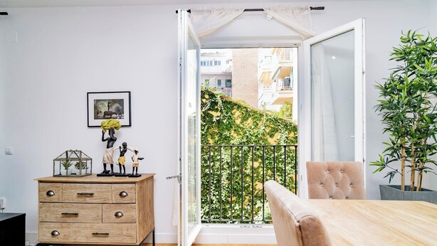 A modern living room with a wooden floor, a beige sofa, a wooden coffee table, a dresser with drawers, a framed picture on the wall, and a balcony with a view of buildings and greenery. - Deluxe Apartment Juan Bravo Suites I (Madrid)