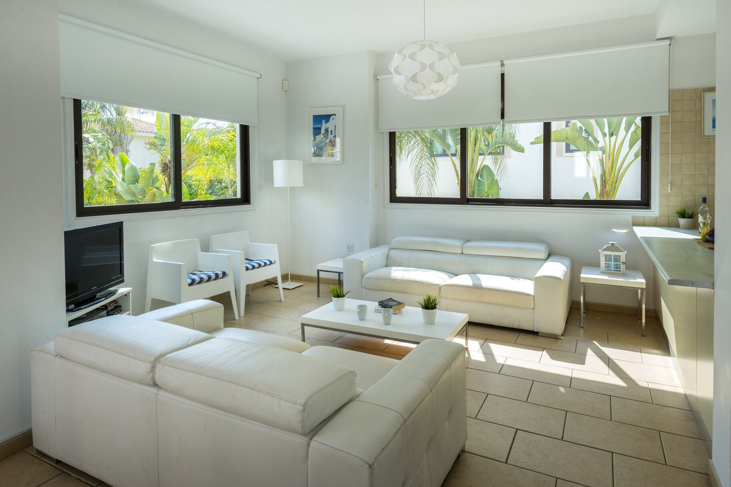 A modern living room with a flat-screen TV, white sofas, a coffee table, and large windows with views of greenery.