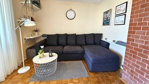 Un salon chaleureux avec un canapé d’angle gris foncé, une table d’appoint ronde avec un pied blanc et un motif géométrique, une horloge blanche accrochée au mur et une petite plante en pot posée sur la table d’appoint.