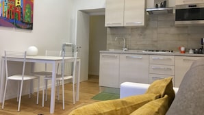 A modern kitchen with white cabinets, a stainless steel range hood, and a microwave. There is a dining area with a white table and chairs, and a colorful painting on the wall. - "G&G Pignasecca" house in the heart of central Naples (Napoli)