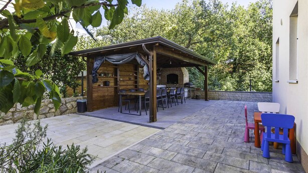 Un pati exterior cobert amb una zona de barbacoa de maó, paviment de pedra i una pèrgola de fusta.