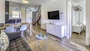 Un saló modern amb un sofà, una taula de menjador, un televisor fixat a la paret i una escala que porta a un altre pis.