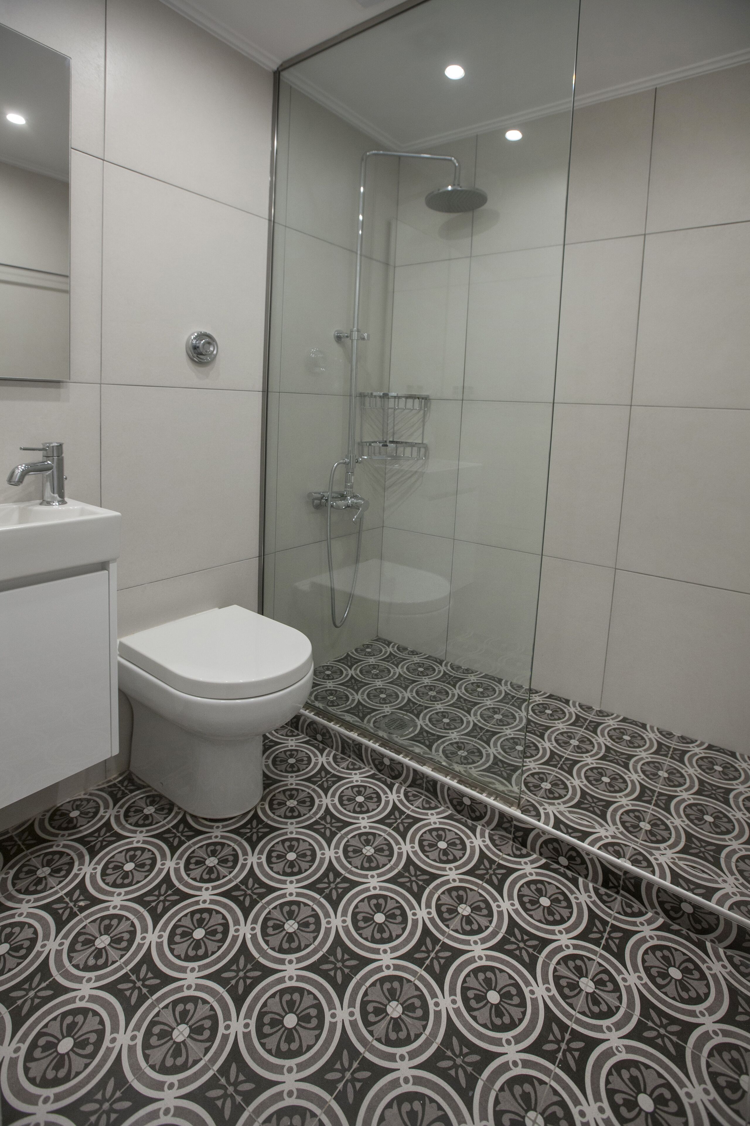 A modern bathroom with a glass-enclosed shower, a white toilet, and a sink. The floor has a patterned tile design.