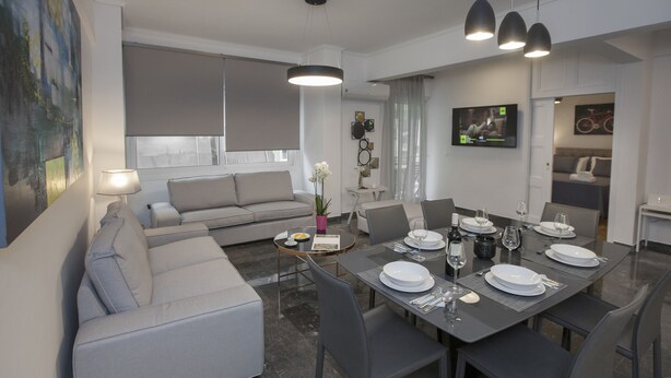 Un salon moderne avec un canapé gris, une table à manger dressée pour quatre personnes et une cuisine en arrière-plan.