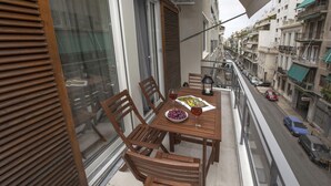 Un balcon agrémenté de meubles en bois, une table avec un saladier de raisins et un verre de vin, et une vue sur la rue en contrebas.