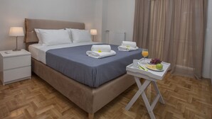 Un lit bien fait, avec du linge de lit blanc et bleu, une table de chevet avec une lampe, et un plateau avec des fruits et un verre de jus d’orange.