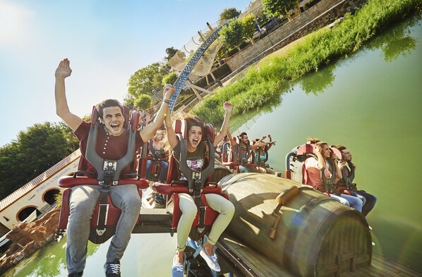 A roller coaster with passengers holding onto the bars, a wooden barrel-shaped structure, and a bridge in the background. - Zeus Attic Tarragona 43005 (Tarragona)