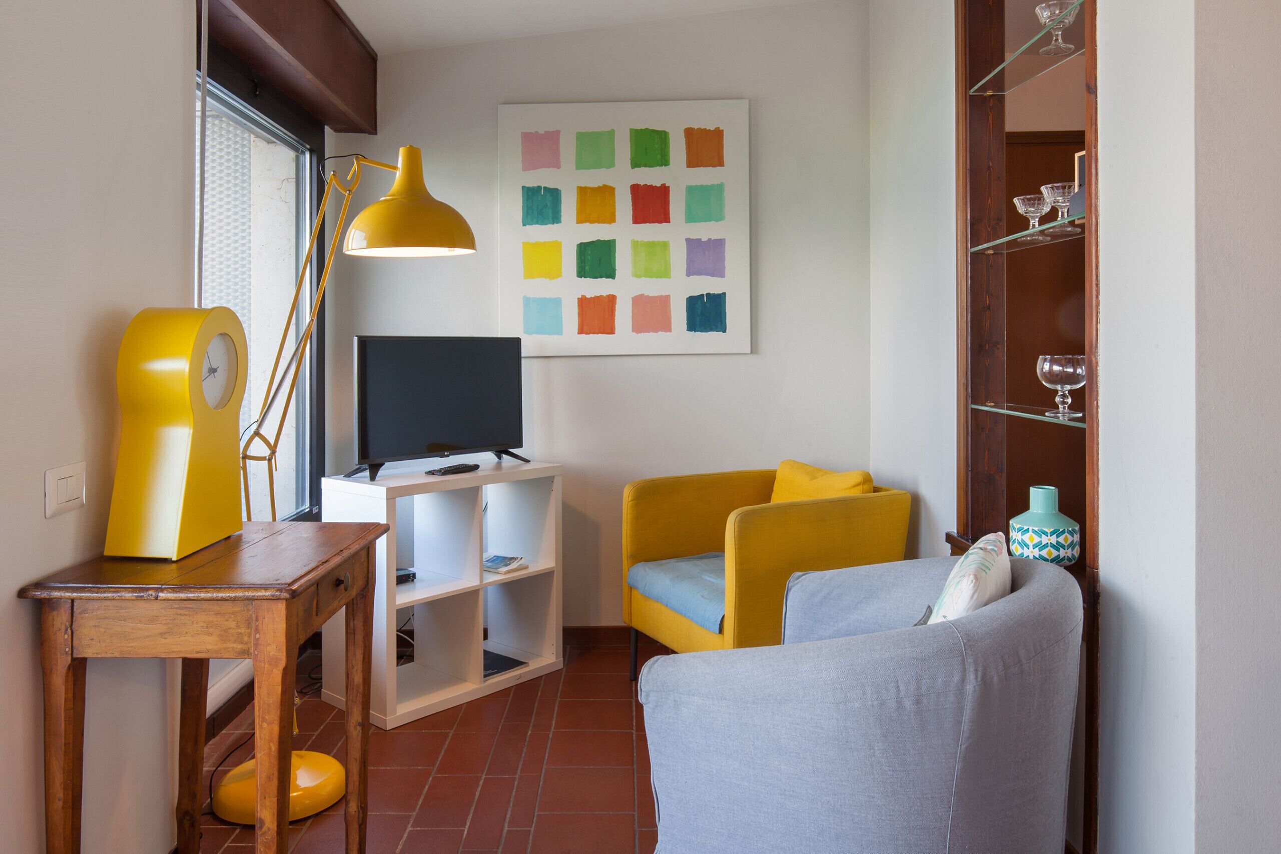 A living room with a TV, yellow and grey sofa, a wooden table, a yellow lamp, and a colorful artwork on the wall.