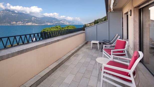 Een balkon met uitzicht op zee en bergen, witte stoelen met rode kussens, en een betegelde vloer.