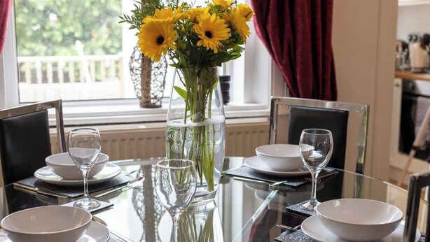 A dining table set with white plates, bowls, and wine glasses, surrounded by chairs. A vase with yellow flowers is placed on the table. - Glebe Street Apartment (Fife)