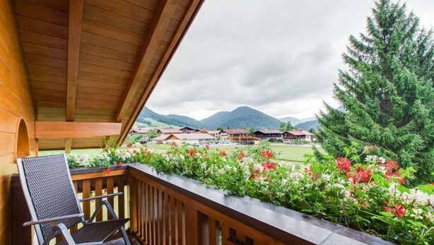 Un balcón de madera con una silla, con vista a un jardín lleno de flores y una vista de casas y montañas.