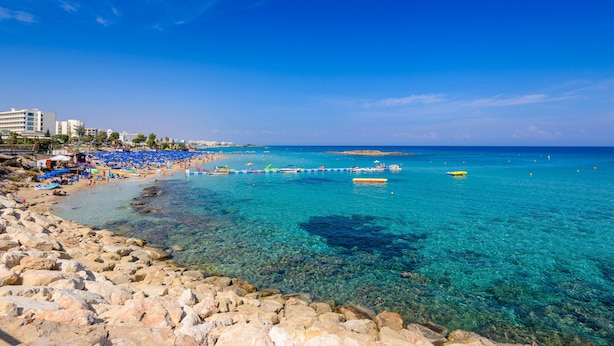 Una playa con agua turquesa cristalina, costa rocosa y sombrillas azules.