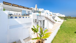 Un edificio moderno de color blanco con un área de estar cubierta al aire libre, una escalera que conduce a un segundo piso y un césped bien cuidado.